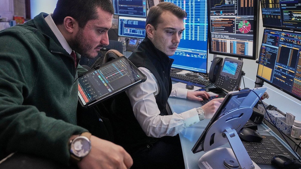 Two traders watch market screens and a tablet at a trading desk